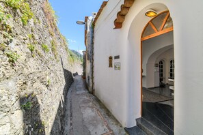 A narrow alleyway with a stone wall on one side and a building entrance with a tiled floor and a door. - Estate4home - Villa Lettieri (Praiano)