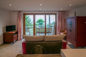 Ein Zimmer mit einer Couch, einem Fernseher, einem Schrank und einer Glastür, die zu einem Außenbereich mit einer Bank und Pflanzen führt.