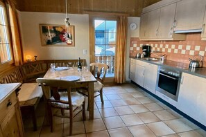 A wooden interior with a dining table, chairs, a kitchen with a stove, and a window with curtains.