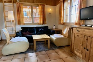 A cozy living room with a black leather sofa, two cream-colored pouf chairs, a wooden coffee table, a TV on a wooden cabinet, and a window with curtains.
