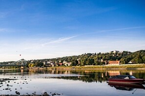 Ein ruhiger Fluss mit einem Boot, das an der rechten Seite vertäut ist, einer Brücke im Hintergrund und einer bewaldeten Anhöhe mit Gebäuden.