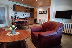 Ein Wohnzimmer mit einem roten Ledersessel, einem Holzesstisch mit Stühlen, einer Küchenzeile mit Holzschränken, einem Heizkörper und einem wandmontierten Fernseher.