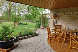 Eine überdachte Terrasse mit Steinboden, Holzmöbeln und Blick auf einen Garten.