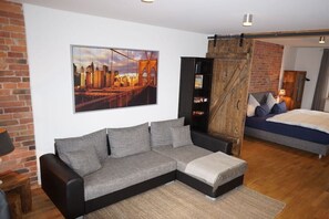 Ein modernes Wohnzimmer mit einem Ecksofa, einem großen Stadtbild, einem Holzschrank und einem Bett mit blauer Kopfteil.
