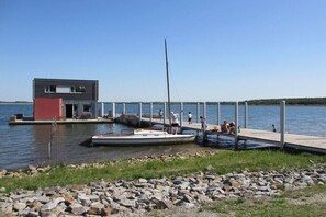 Ein modernes Gebäude mit einer Anlegestelle, die sich in ruhiges Wasser erstreckt, ein kleines Boot liegt an der Anlegestelle festgemacht, und Personen befinden sich in der Nähe.