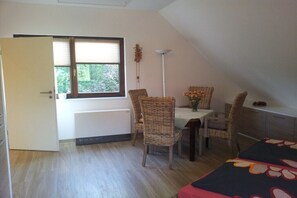 Ein Zimmer mit Esstisch, Korbstühlen, Heizkörper, Stehlampe und einem Bett mit Blumenmuster-Zudecke.