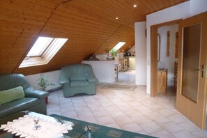 Ein Loft-Zimmer mit schrägverdeckter Decke, einem grünen Sofa, einem Esstisch und einer Küchenzeile im Hintergrund.