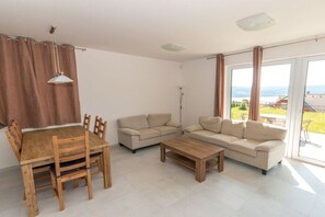 Ein Wohnzimmer mit einem Holzesstisch, beigen Sofas, einem Couchtisch und großen Fenstern mit Blick ins Freie.