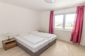 Ein Zimmer mit zwei Betten, einem Nachttisch, einem Fenster mit Vorhängen und Blick auf Gebäude.
