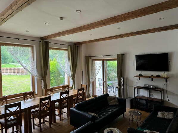 A living room with a dining area, wooden beams, a TV, and a large window.