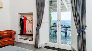 Una habitación con una butaca de cuero marrón, un perchero con diversas prendas y un ventanal corredero que da a un patio exterior con una mesa y sillas.