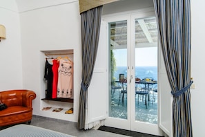 A room with a brown leather chair, a clothing rack with various garments, and a sliding glass door leading to an outdoor patio with a table and chairs. - Estate4home - Villa Lettieri (Praiano)