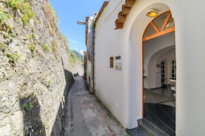 A narrow alleyway with a stone wall on one side and a white building with an arched entrance on the other. - Estate4home - Villa Lettieri (Praiano)