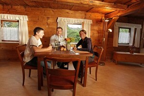 Interior de uma cabana de madeira com mesa de jantar, cadeiras e uma janela com cortinas.