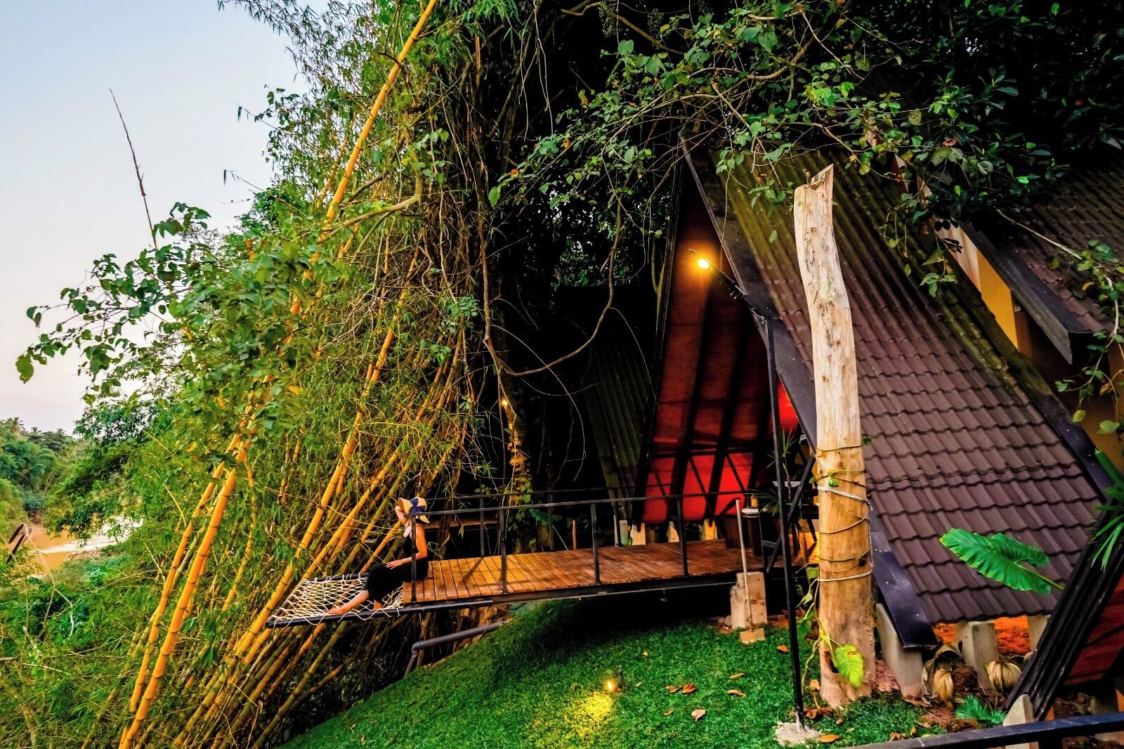 A rustic wooden structure with a sloped roof, surrounded by dense foliage and a small wooden bridge.