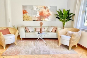 Ein Wohnzimmer mit einer Couch, zwei Korbstühlen, einem Glastisch, einem großen abstrakten Gemälde und einer üppigen Grünpflanze.