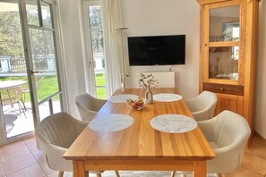Ein Essbereich mit einem Holztisch, weißen Stühlen, einer Vase mit Blumen und einem an der Wand befestigten Fernseher.