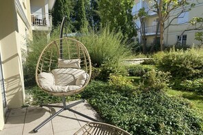 Ein Korbstuhl mit Kissen in einem Garten mit üppiger Bepflanzung und einem Gebäude im Hintergrund.