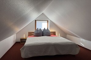 Ein Schlafzimmer mit schrägen Wänden, einem Bett mit weißer Bettdecke, zwei Nachttischen mit Lampen und einem Fenster mit Jalousien.