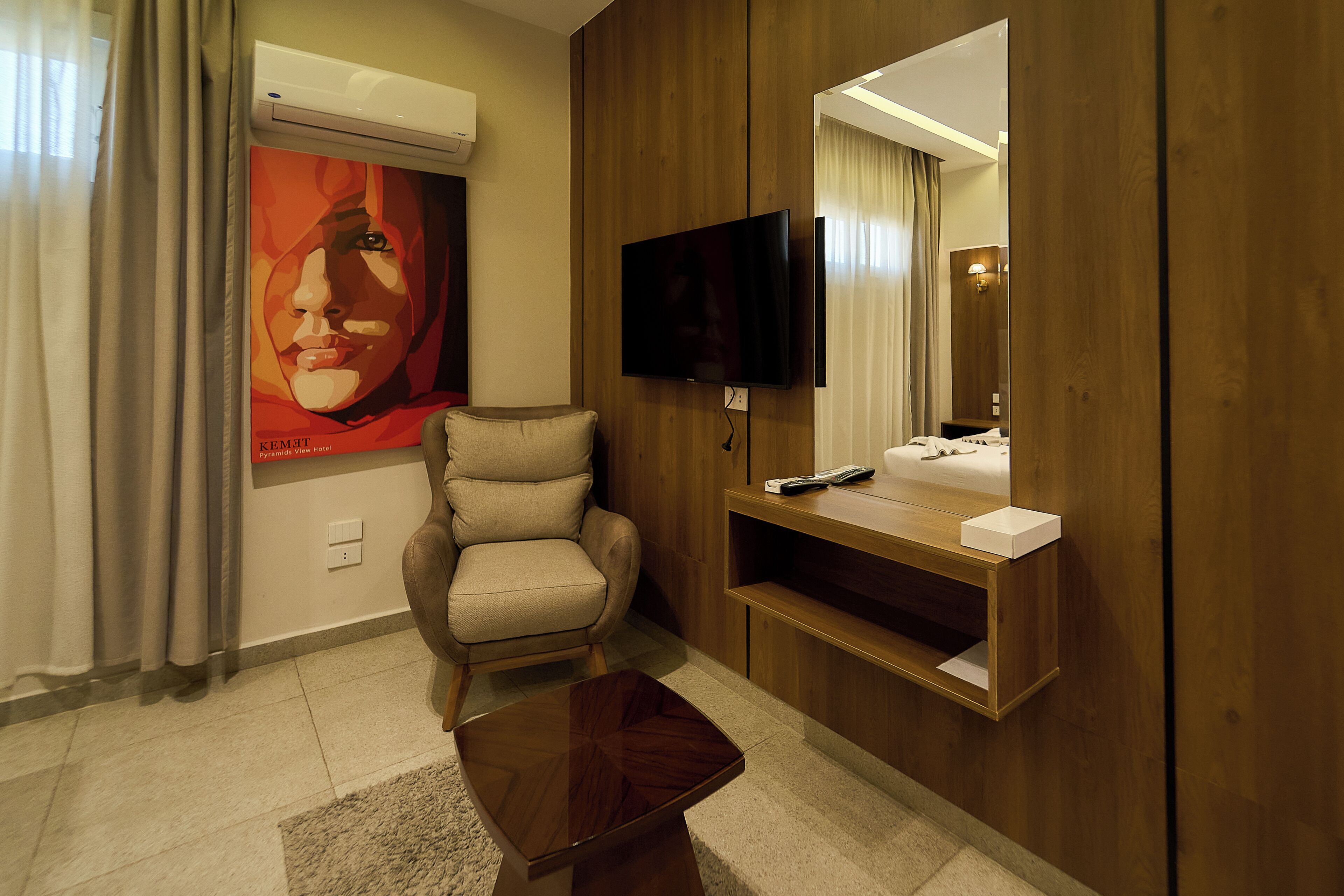 A hotel room with a flat-screen TV mounted on the wall, a wooden vanity with a mirror, a beige armchair, and a small coffee table.