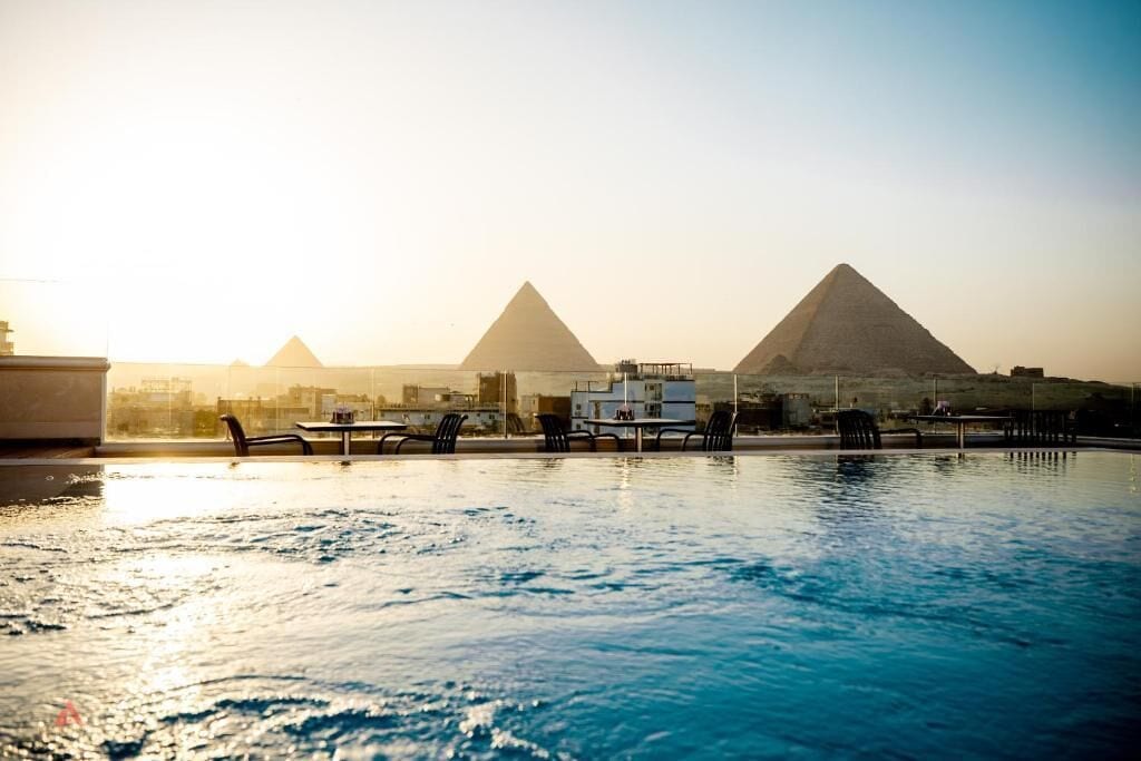 A swimming pool with lounge chairs and tables, with the pyramids in the background.