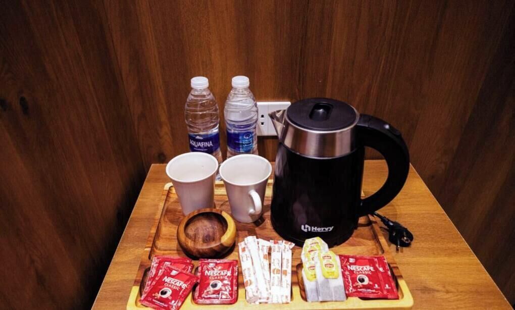 A wooden table with a black coffee maker, two water bottles, two cups, a bowl, and several packets of Nescafé Classic instant coffee.