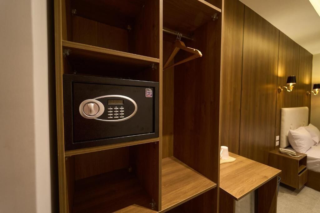 A hotel room with a built-in safe, wooden wardrobe, a bedside table with a lamp, and a bed with white linens.