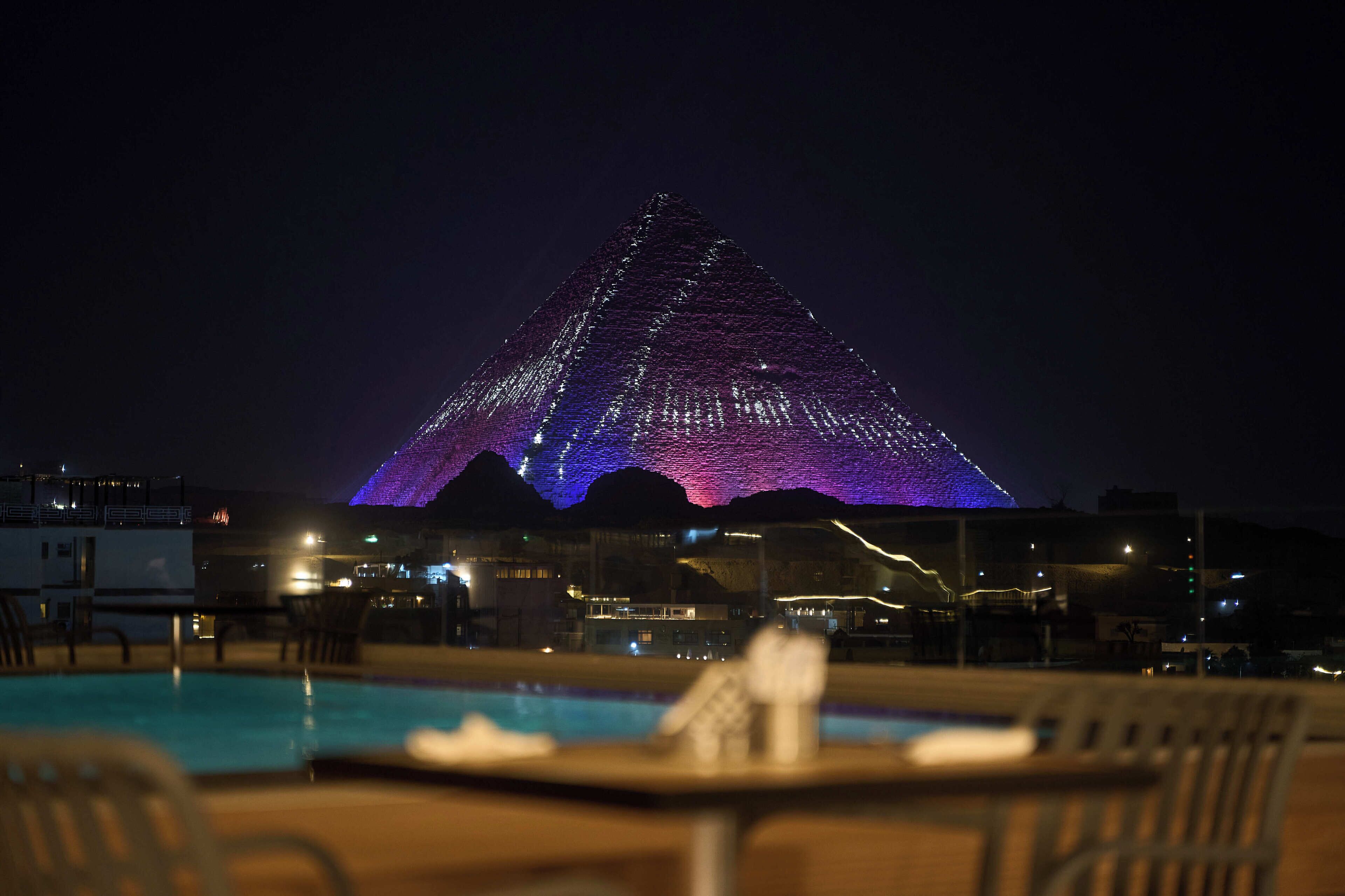 A pool area with a view of a pyramid-shaped building illuminated with purple and blue lights.