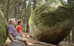 Deux personnes sont assises sur un rocher, dans une zone boisée, près d’un gros bloc de pierre recouvert de mousse.