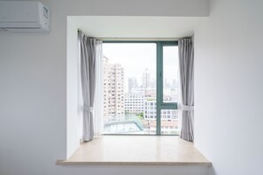 A room with a large window, a view of high-rise buildings, and an air conditioning unit mounted on the wall.