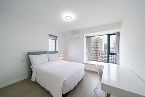 Ein Schlafzimmer mit einem Bett, einem Schreibtisch, einem Fenster mit Blick auf Gebäude, einer Klimaanlage und einer Deckenleuchte.