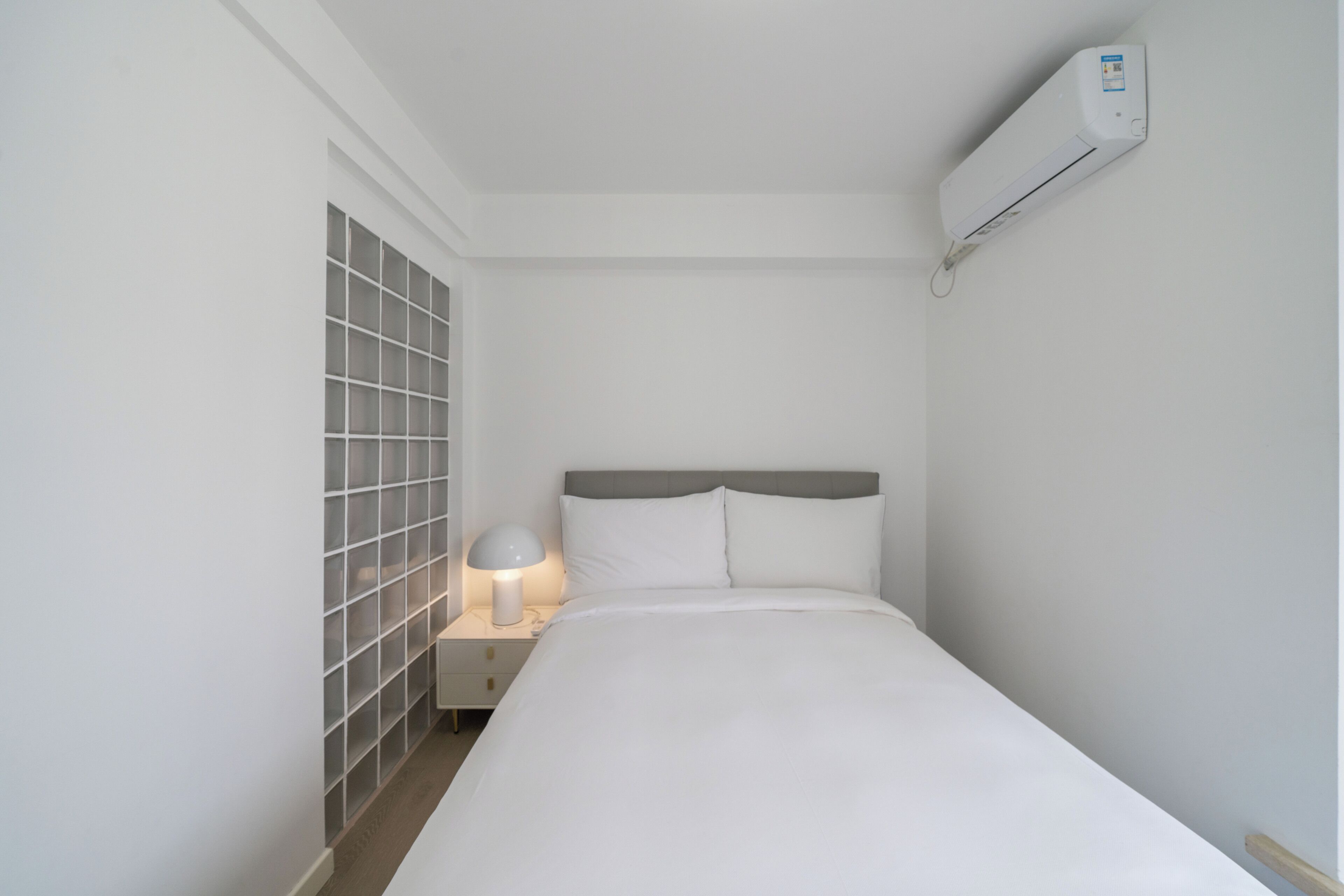 A bedroom with a bed, a nightstand with a lamp, a wall-mounted air conditioner, and a window with blinds.