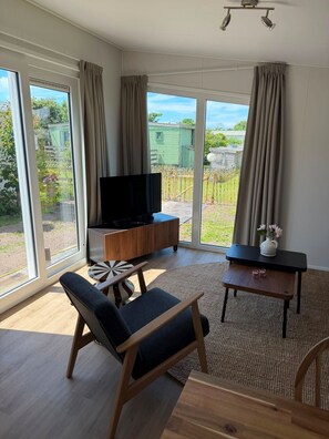 Ein modernes Wohnzimmer mit Fernseher, Couchtisch aus Holz, gepolstertem Sessel und großen Fenstern mit Blick auf einen Garten und ein grünes Gebäude.