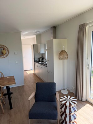 Ein modernes Interieur mit Essbereich, einem blauen Stuhl, einer Küche mit weißen Schränken und einem großen Fenster mit Vorhängen.
