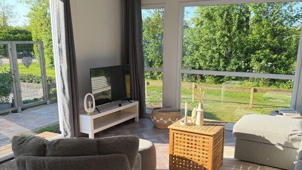 A modern living room with a grey sofa, a TV on a white stand, a wooden side table, and a view of a green garden through large windows. - Chalet 193 (for 4 persons) (Petten)