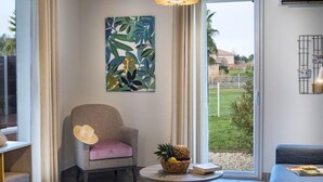 A room with a chair, a table with a book and a fruit basket, a painting on the wall, and a view of the outdoors. - French Villa with Shared Pool (Fabrègues)