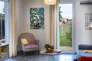 A room with a chair, a table with a book and a fruit basket, a painting on the wall, and a view of the outdoors. - French Villa with Shared Pool (Fabrègues)