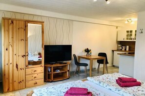 Ein Zimmer mit Holzschrank, Fernseher, Schreibtisch mit Stuhl, Bett mit floralem Bettbezug und einer Küchenzeile.