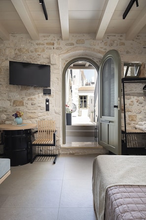A room with a stone wall, a bed, a TV mounted on the wall, a chair, and a small table with a vase. - L10 Old Town Boutique Studios (Rethmyno)