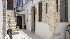 Un patio con un muro de piedra, una escalera, un balcón con mobiliario exterior y una puerta que da a un espacio interior.