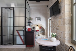 A room with a stone wall, a fireplace, a round table, and a television mounted on the wall. - L10 Old Town Boutique Studios (Rethmyno)