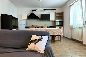 A living room with a sofa, a table with chairs, a kitchen area with appliances, and a window with curtains. - Location: in the central area, a few steps from the sea and all services.Description: very central four-room apartment on two floors connected by stairs.1st floor: large living room with sofa, table, kitchenette and dishwasher - bedroom with double bed - (Cesenatico)
