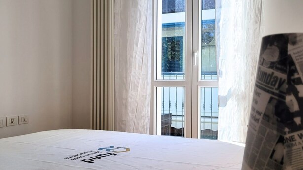 A hotel room with a large window, a bed with a white bedspread, and a partial view of a newspaper. - Location: in the central area, a few steps from the sea and all services.Description: very central four-room apartment on two floors connected by stairs.1st floor: large living room with sofa, table, kitchenette and dishwasher - bedroom with double bed - (Cesenatico)