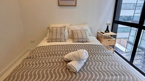 En pænt redt seng med et mønstret sengetæppe, et sengebord med lampe, et billede i ramme på væggen og et stort vindue med udsigt til bygninger.