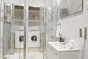 Ein modernes Badezimmer mit einer gläsernen Duschkabine, einer integrierten Waschmaschine und einem weißen Waschbecken mit Spiegel.