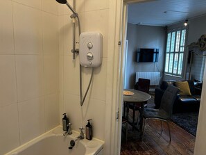 Une salle de bain avec une douche, une baignoire et des toilettes. On aperçoit, à travers l’embrasure de la porte, un coin repas avec une table et des chaises.