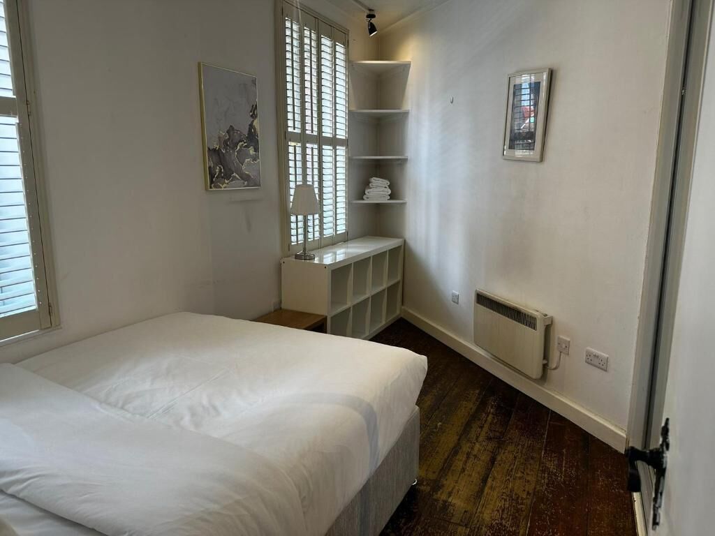 Un dormitorio con cama, ventana con persianas, radiador y estantería.