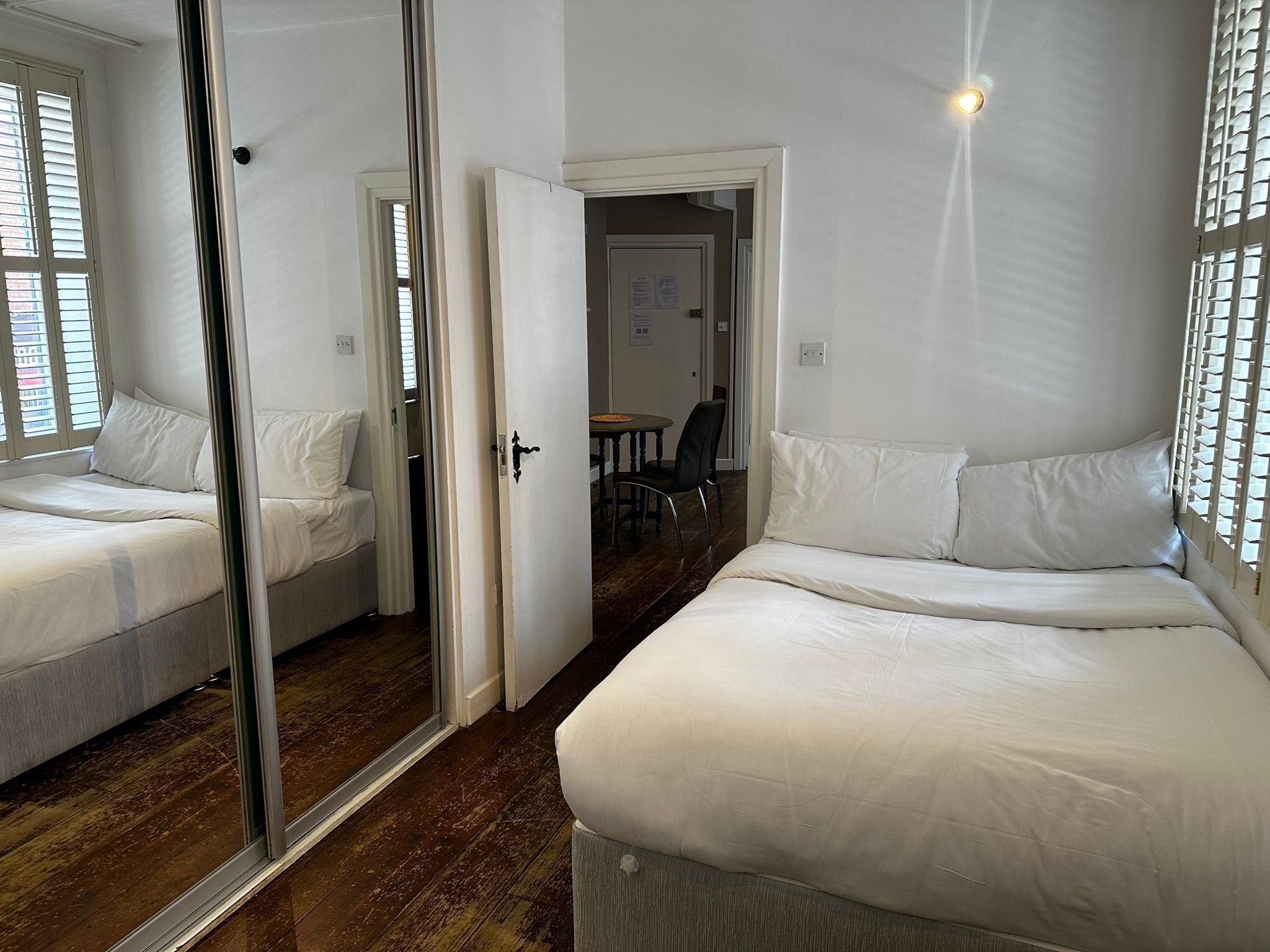Une chambre d’hôtel avec deux lits, un miroir, une porte donnant sur un coin repas et une fenêtre avec des stores.