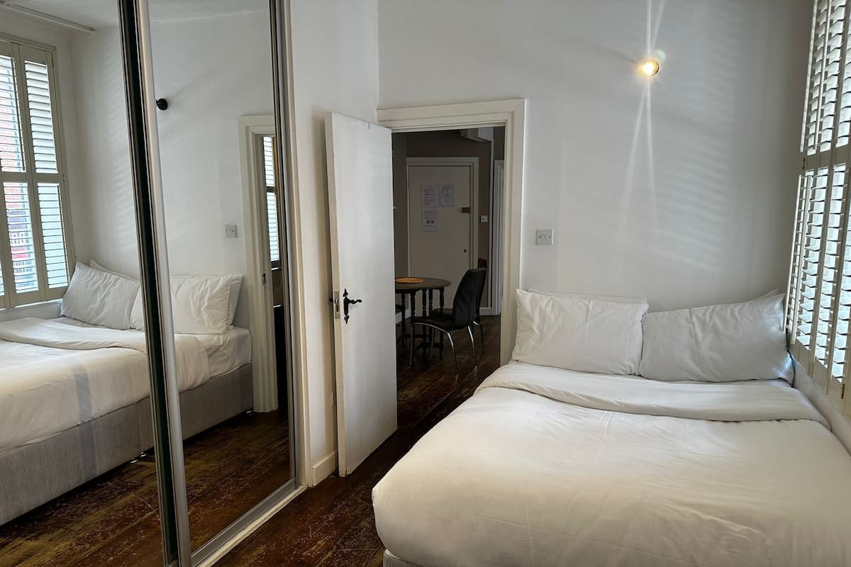 Quarto de hotel com duas camas, um espelho, uma porta que dá para uma sala de jantar e uma janela com persianas.