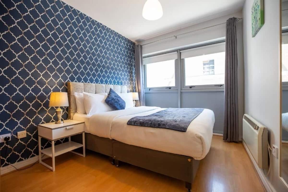 Quarto com parede estampada, cama com almofadas, mesa de cabeceira com abajur e janela com cortinas.